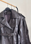 A dark brown textured leather trench coat hanging on a black hanger from a wooden clothes rack against a plain off-white wall.